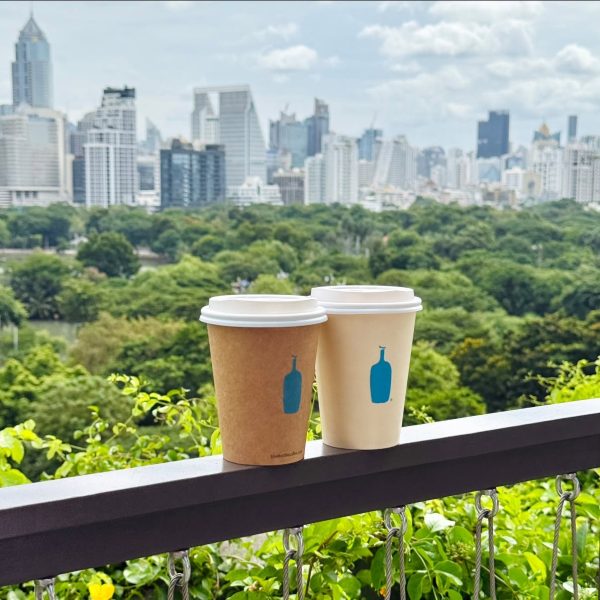 แก้วกาแฟ Blue Bottle สองแก้ววางบนระเบียงด้านนอก เบื้องหลังเป็นวิวสวนสีเขียวและตึกสูงใจกลางเมือง