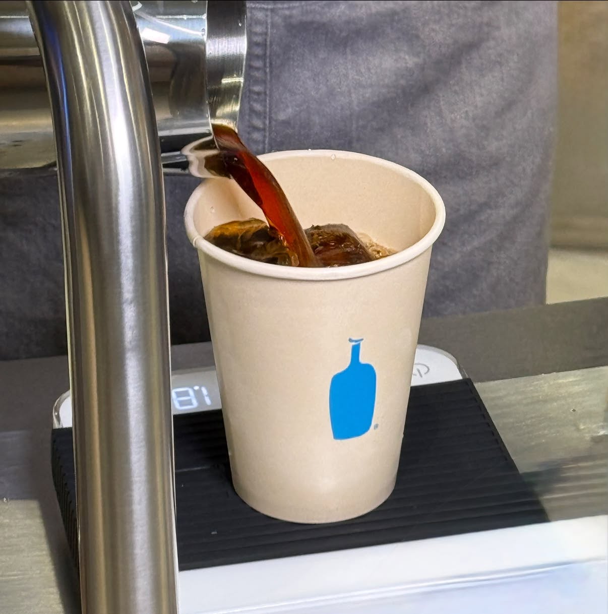 ภาพระยะใกล้ขณะกำลังรินกาแฟ Cold Brew ลงในแก้วกระดาษ Blue Bottle บนเครื่องชั่งดิจิทัล